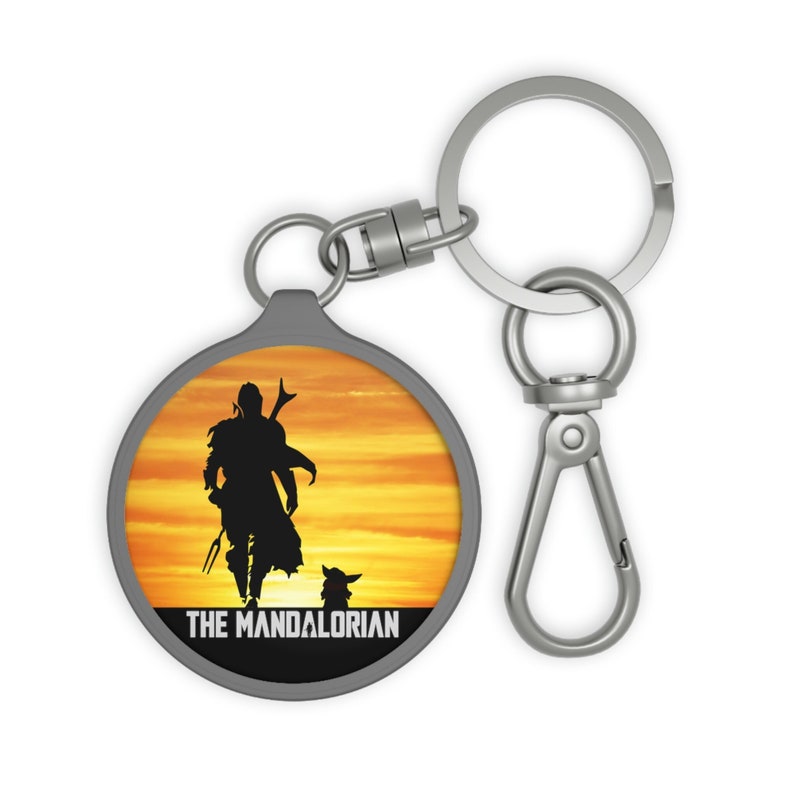 May include: A silver keychain with a round, metal charm featuring a silhouette of The Mandalorian and Grogu walking into a sunset. The text "THE MANDALORIAN" is printed below the image.