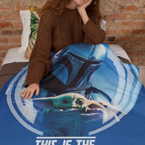 May include: A blue throw blanket with a graphic of a Mandalorian character holding a small green alien. The text "This is the Way" is printed in white on the blanket.