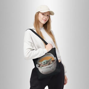 Puede incluir: Una bolsa de tela negra con una correa blanca. La bolsa presenta un gr&aacute;fico de un personaje alien&iacute;gena verde sentado en una c&aacute;psula redonda. El alien&iacute;gena lleva una t&uacute;nica marr&oacute;n y tiene orejas grandes.