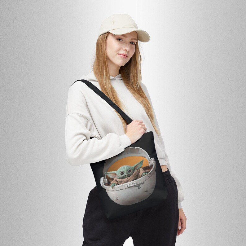 Puede incluir: Una bolsa de tela negra con una correa blanca. La bolsa presenta un gr&aacute;fico de un personaje alien&iacute;gena verde sentado en una c&aacute;psula redonda. El alien&iacute;gena lleva una t&uacute;nica marr&oacute;n y tiene orejas grandes.