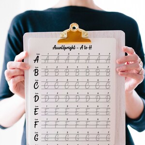 May include: A white clipboard with a black and white handwriting practice sheet for the letters A through G. The sheet has lines and numbers to guide the user. The title of the sheet is "AsunUpright - A to H".
