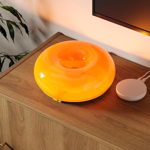 May include: An orange glass lamp with a circular design sits on a wooden cabinet. A white circular device and a stack of books are next to the lamp. A black game controller rests on top of the books. The cabinet is in front of a black television.