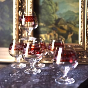 Può includere: Un gruppo di bicchieri da brandy in vetro trasparente con fasce decorative rosse e dorate. I bicchieri sono disposti su una superficie scura, con un dipinto incorniciato sullo sfondo. Ogni bicchiere contiene circa 120-180 ml.