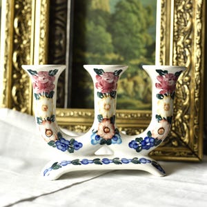 May include: A white ceramic candelabra with three candle holders, adorned with hand-painted floral designs in pink, blue, and yellow. The decorative piece is set against a blurred background with a gold framed painting.