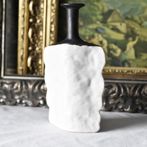 May include: A white ceramic vase with a textured surface and a black neck. The vase has a rectangular shape with rounded edges. The background includes a framed painting and a gold-coloured frame.