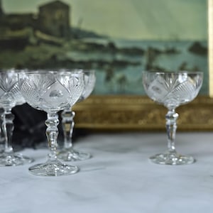 May include: Several clear crystal coupe glasses with detailed etched patterns. The glasses have a stemmed base and a broad, shallow bowl. A framed painting with a gold border is in the background.