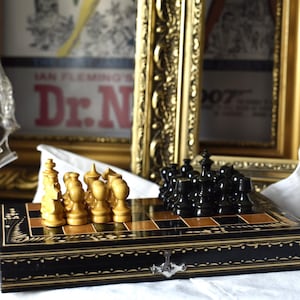Può includere: Un set di scacchi vintage con pezzi neri e color crema su una scacchiera intarsiata in nero e oro. La scacchiera è appoggiata su un panno bianco, con un poster incorniciato sullo sfondo.
