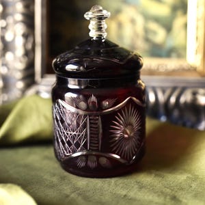 Könnte beinhalten: Ein rubinrotes Glasgefäß mit Deckel, das aufwendige geätzte Designs aufweist. Das Gefäß hat eine runde Form mit einem detaillierten Muster aus Linien und Sternen. Der Deckel ist mit einem transparenten, dekorativen Knopf versehen. Das Gefäß steht auf einer grünen Oberfläche.