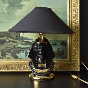 Puede incluir: Una lámpara de mesa negra con una pantalla de tela negra y una base dorada. La base presenta un busto esculpido de un faraón egipcio.