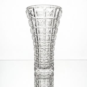Op de afbeelding: Een heldere glazen vaas met een herhalend geometrisch rasterpatroon van verhoogde vierkanten over het gehele buitenoppervlak. De vaas heeft een uitlopende opening aan de bovenkant en loopt iets taps toe naar de basis, met een transparante afwerking.