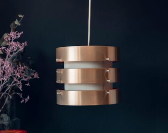 May include: A copper-colored pendant light with three tiers hangs from a white cord against a dark blue background. The light has a white diffuser in the center.