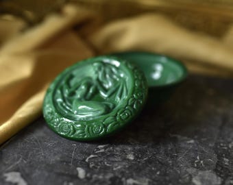 May include: A green glass trinket box with a raised design of a woman surrounded by roses. The box is closed and sitting on a dark surface.