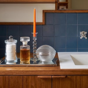 May include: A rectangular silver mirrored tray holding a clear glass canister filled with white cotton rounds, a ribbed glass decanter with amber liquid, a tall clear glass candle holder with an orange taper candle, and a round textured glass bowl.