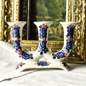 May include: A white ceramic candelabra with three candle holders. The candelabra is decorated with blue and pink floral patterns. The background includes a framed painting and a gold-colored frame.