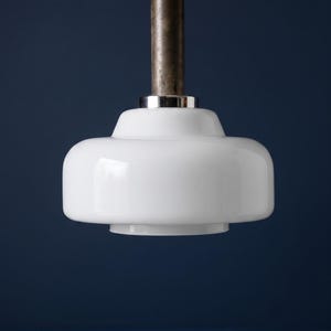 Könnte beinhalten: Eine Pendelleuchte mit einem glatten, weißen, abgerundeten Glasschirm, der an einem zylindrischen, dunkelmetallischen Schaft befestigt ist. Der Schaft ist oben mit einem schwarzen Kabel verbunden, und ein polierter silberfarbener Metallring sitzt zwischen Glas und Schaft.