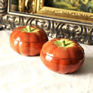 以下が含まれることがあります： リンゴの形をした2つのセラミック製の小物入れ。箱は光沢のある赤みがかったオレンジ色で、緑色の茎が付いています。白い布の上に置かれ、背景には装飾的な額入りの絵画があります。