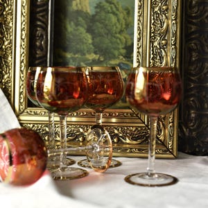 Peut inclure: Ensemble de verres à vin vintage avec une teinte rouge-orangé et des bords dorés. Les verres sont disposés devant une peinture encadrée avec un cadre doré orné. La scène se déroule sur une surface blanche.