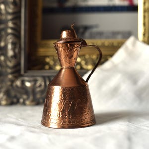 Könnte beinhalten: Eine kupferfarbene dekorative Kanne mit Griff und Deckel. Die Kanne hat ein verziertes, geprägtes Design und steht auf einer weißen Oberfläche. Der Hintergrund enthält ein gerahmtes Bild.