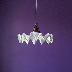 Peut inclure: Une suspension en verre blanc avec un bord ondulé et plissé souligné d'un liseré vert. La lampe est dotée d'une douille en métal foncé et est suspendue par un fin cordon argenté. L'abat-jour présente un design plissé avec un bord festonné.