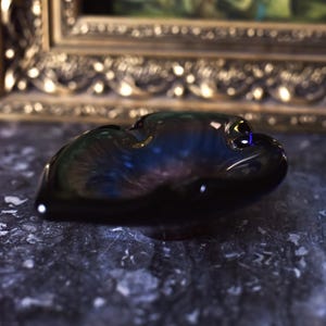 May include: A dark, glossy, black glass ashtray with an unusual, organic form. The ashtray reflects light, showing hints of blue and purple. The background includes a framed painting with gold detailing.