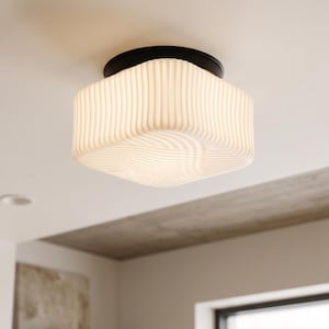 May include: A flush mount ceiling light fitting featuring a square, white, ribbed glass shade with rounded corners. The shade is attached to the ceiling by a circular, matte black metal base, providing a clean and functional lighting solution.