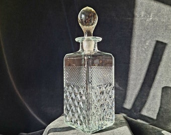 Puede incluir: Una decantador de vidrio transparente con un patrón de diamante y un tapón de vidrio. La decantador está sobre una superficie de tela gris.