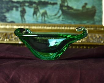 May include: A striking green glass bowl with a distinctive curved form. The bowl, with its glossy surface, is set against a dark red backdrop. The edges of the bowl curve upwards, creating a unique shape.