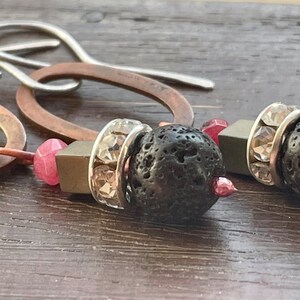 May include: A pair of earrings with copper oval hoops, silver hooks, and black lava rock beads. The beads are accented with clear rhinestones and pink gemstone accents.