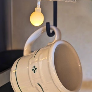 May include: A black Keurig coffee maker with a white ceramic mug hanging from a hook. The mug has a green floral design and is partially filled with coffee. String lights are visible in the background.