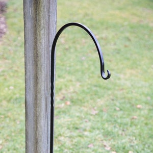 Puede incluir: Gancho de metal negro fijado a un poste de madera. El gancho es curvo y tiene un giro decorativo en la base.