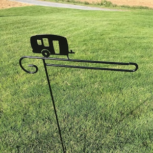Puede incluir: Soporte de bandera de jardín de metal negro con una silueta de una caravana en la parte superior. El soporte está en un césped verde.