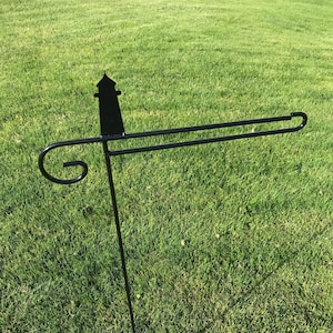 May include: Black metal garden flag stand with a lighthouse silhouette at the top. The stand has a curved arm and a pointed base.