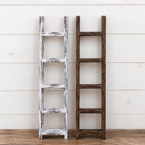 May include: Two decorative wooden ladders standing upright. One ladder features a distressed white paint finish over dark wood, while the other ladder has a solid, dark brown stained wood finish. Both ladders consist of four horizontal rungs.