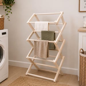 May include: A wooden clothes airer with three towels in white, green, and beige. The airer is in a laundry room with a washing machine and a wicker laundry basket. The rack is made of natural wood.