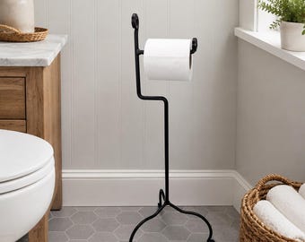 May include: A black metal freestanding toilet paper holder with a scroll-style base and a single horizontal arm that holds one roll of white toilet paper. The stand features a slender vertical post and a decorative curled top detail.