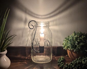 May include: A clear glass mason jar lantern featuring a black metal scroll handle and a central black candle holder. A white taper candle is lit inside the jar, casting a warm glow and distinct light patterns against the wall behind it.
