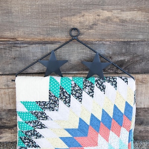 May include: Black metal quilt rack with two star-shaped hooks hanging on a wooden wall. A colorful patchwork quilt is hanging on the rack.