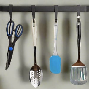 May include: A black metal kitchen utensil rack with four hanging utensils. Utensils include scissors with blue accents, a slotted spoon, a blue spatula, and a slotted turner. The rack is mounted on a light gray wall.