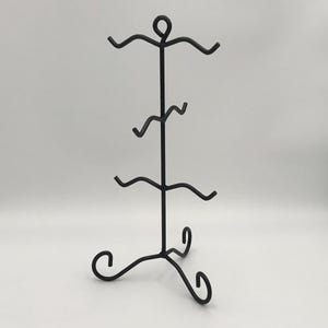 May include: A black metal jewellery display stand with a three-legged base and a vertical post. The post has four curved arms to hold items. The top of the stand has a circular design. The stand is designed to hold jewellery.