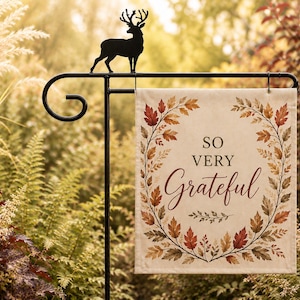 May include: A rectangular garden flag hanging from a black metal stand topped with a deer silhouette. The flag features a cream background with a wreath of autumn-colored leaves in shades of red, orange, and brown, and the text "SO VERY Grateful".