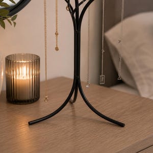 May include: A black metal jewelry tree displaying several necklaces with gold and silver chains. A lit candle in a ribbed glass holder and a decorative vase with greenery sit on a wooden surface. A mirror and a bed are in the background.