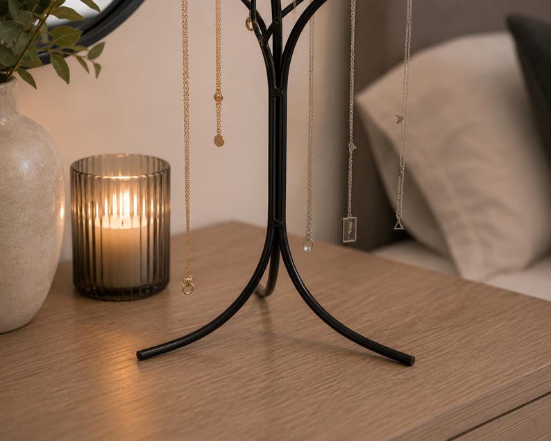 May include: A black metal jewelry tree displaying several necklaces with gold and silver chains. A lit candle in a ribbed glass holder and a decorative vase with greenery sit on a wooden surface. A mirror and a bed are in the background.