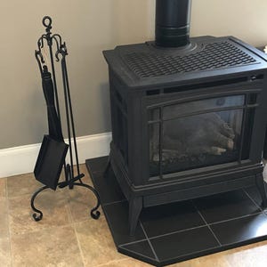 Puede incluir: Estufa de leña de hierro fundido negro con una puerta de vidrio y una rejilla de metal negro en la parte superior. La estufa está sobre un suelo de baldosas negras. Un juego de herramientas para chimenea de metal negro se encuentra en la esquina de la imagen.