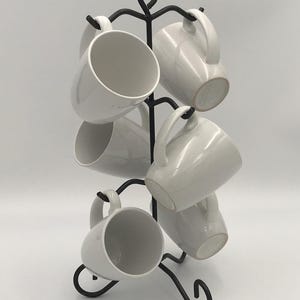 May include: A black metal mug tree displaying six white ceramic mugs. The mugs have a simple design with a curved handle. The mug tree has a decorative, swirling design at the top and base.