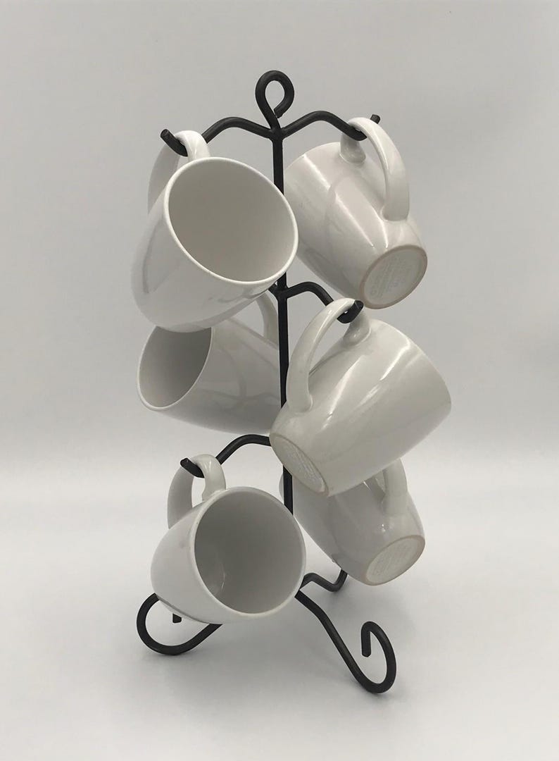 May include: A black metal mug tree displaying six white ceramic mugs. The mugs have a simple design with a curved handle. The mug tree has a decorative, swirling design at the top and base.