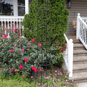 May include: Black metal garden trellis with a pointed top and multiple tiers. The trellis is standing in a garden bed with pink flowers.