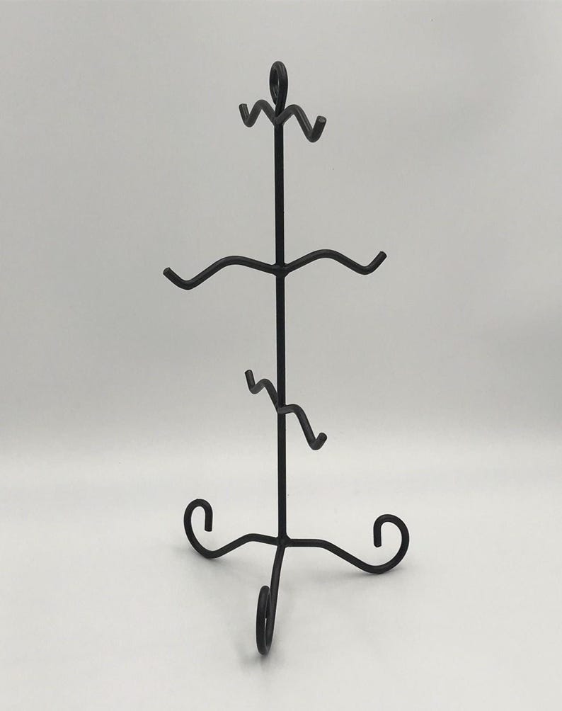 May include: Black metal display stand with a three-tiered design. The stand has decorative scrollwork at the base and top, with curved arms to hold items. Suitable for displaying jewellery, small art, or collectibles.