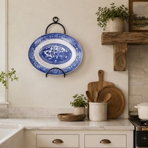 May include: An oval blue and white decorative plate with a detailed pattern is displayed on a black metal plate holder. The plate is mounted on a cream-colored wall in a kitchen setting. The kitchen has a farmhouse style.