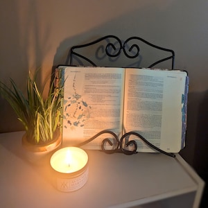 Puede incluir: Una vela encendida con una etiqueta blanca que dice "Marcus Harvey" se encuentra sobre una mesa junto a un libro cerrado con un soporte de libro de metal negro. El libro tiene una cubierta azul y blanca con un diseño de peces y las palabras "Jesús es la paz de mi alma".