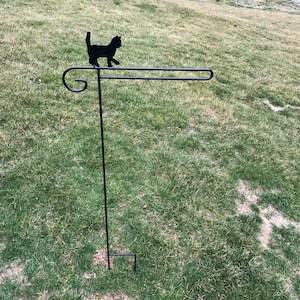 May include: Black metal garden stake with a silhouette of a cat walking on a horizontal bar. The stake is inserted into a grassy lawn.